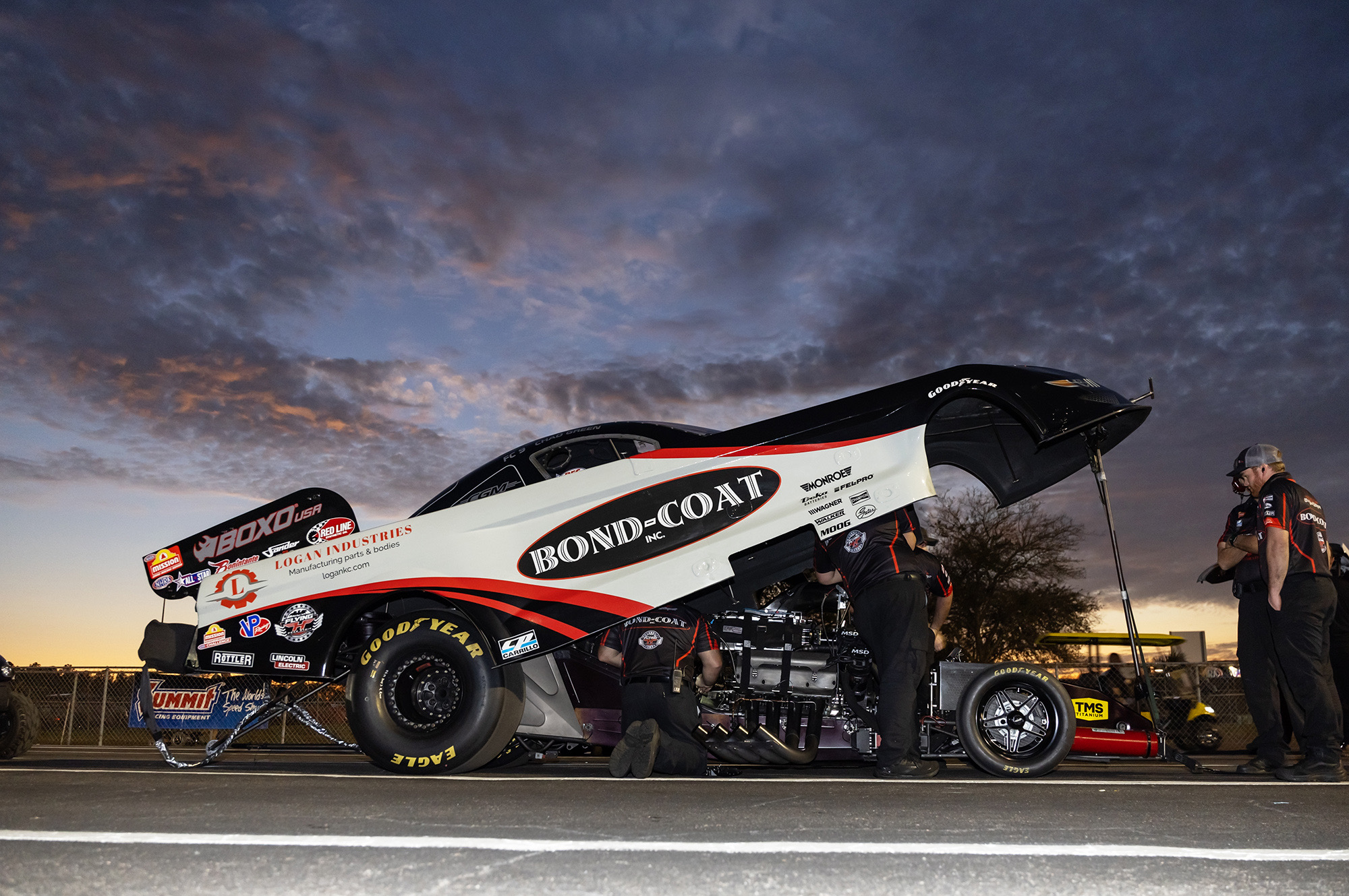 The Bond-Coat Funny Car sits with its body raised at dusk as the crew works beneath a storm sky